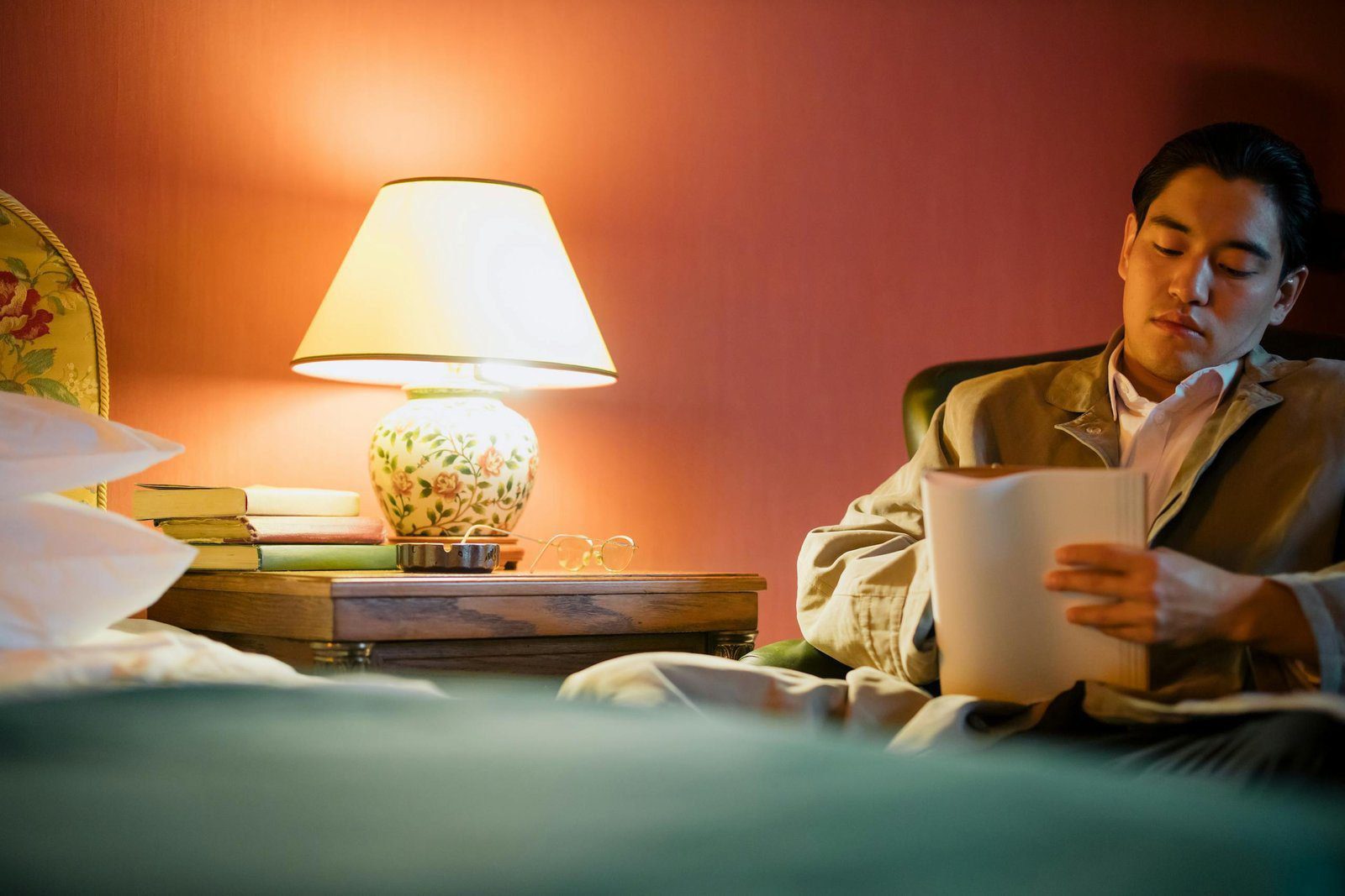 An adult man reads in a warmly lit hotel bedroom with a lamp and bedside table.